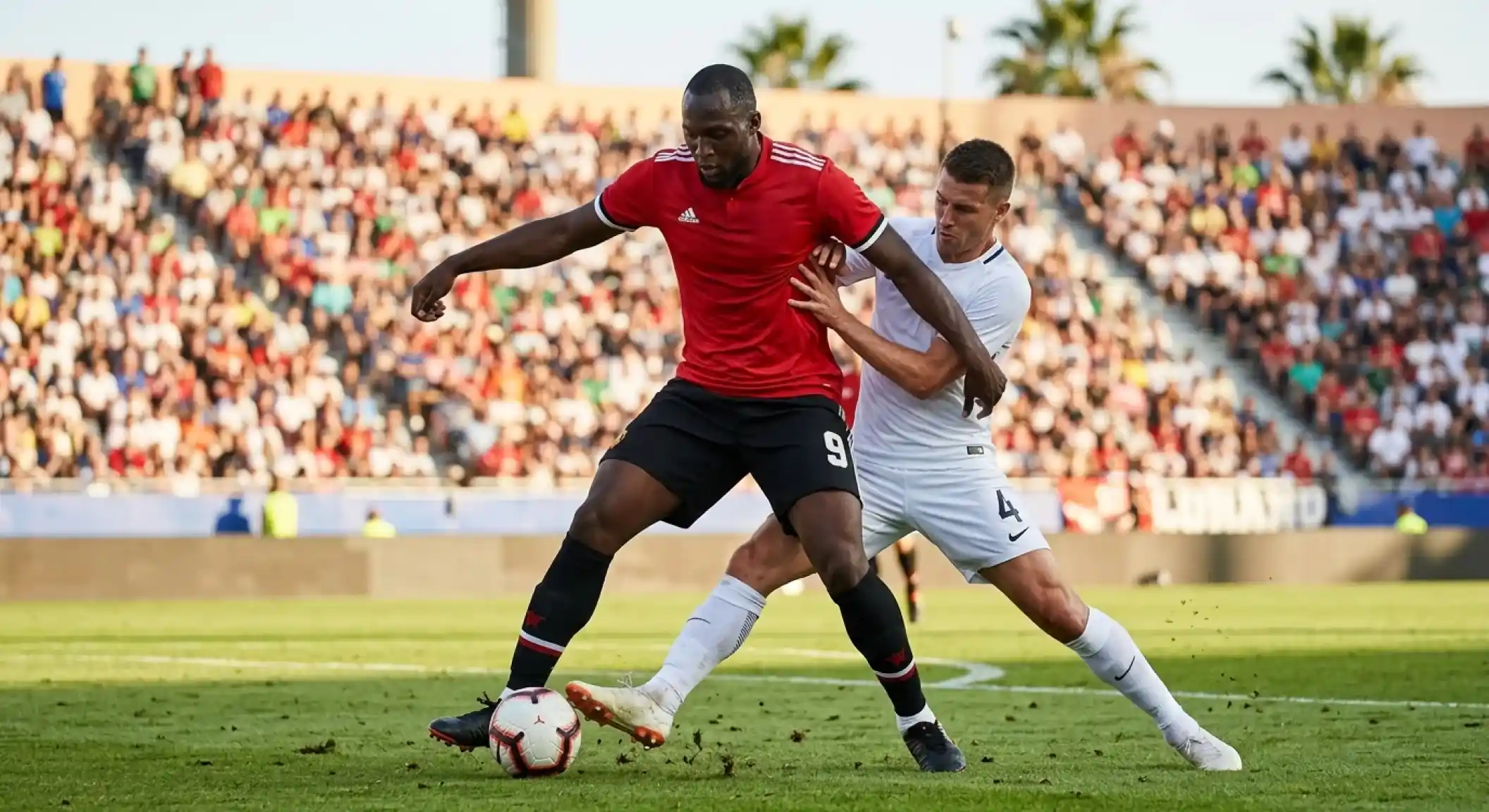 A tall striker in a red kit shielding the ball from a defender in white under bright sunshine at an open stadium, intense knockout duel
